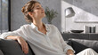 © Vyatcheslav - Young woman relaxing on sofa with eyes closed in modern living room. Happy female resting at home in white shirt. Wellness and comfort concept