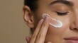 © Viktor - Young woman applies white moisturizer cream on cheek for daily skincare routine. Beautiful female model touches face with fingers to massage cosmetic product. Gentle lady cleanses, hydrates healthy