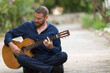 © Fatih - Portrait of a middle-aged bearded man playing acoustic classical guitar, sitting cross-legged outdoors in a garden with natural bokeh background.