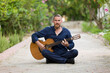 © Fatih - Portrait of a middle-aged bearded man playing acoustic classical guitar, sitting cross-legged outdoors in a garden with natural bokeh background.