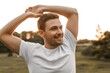 © Lamia - Handsome young man with a beard wearing a plain white t-shirt stretching his arms overhead while enjoying a peaceful moment in a park during a warm golden hour sunset