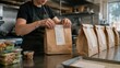 © olga_demina - Hands of food service worker sealing labeled paper takeaway bag on stainless counter amid lined row of prepared orders, with salad containers and condiment cup visible in foreground and industrial