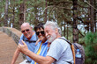 © luciano - Three smiling senior friends hiking in a green pine forest, capturing a joyful moment together with a smartphone, actively enjoying nature and companionship