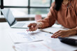 © David - Professional woman analyzing financial data and business reports, using a pen to point at charts while working on a laptop at an office desk