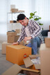 © Pichsakul - Young Asian man sealing cardboard box with packing tape while unpacking belongings in bright modern apartment