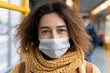 © lashkhidzetim - Woman wears protective mask on public transport. She looks directly at camera with calm, thoughtful gaze. Cozy knitted scarf wraps her neck, adds warmth