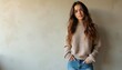 © Maryna - Young woman in beige sweater and jeans stands with hands in pockets. She has long brown hair and a confident, relaxed expression against a textured neutral wall.