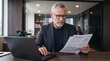 © Eve - Senior businessman wearing glasses analyzing financial charts document paper at office desk with laptop mature corporate executive leader managing investment data accounting strategy plan