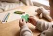 © Olena Apetroaiei - Child holds a green heart cutout while sitting at a wooden table with art supplies in a cozy indoor space
