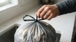 © SimpleDesignStudio - Adult male hand holding a tied grey plastic trash bag in a kitchen, representing household waste management and daily chores
