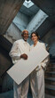 © f_bossa - Two people stand in concrete staircase enclosure while holding a large white rectangular board. Soft daylight filters through overhead window onto their pale cream clothing.