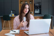© simona - Young woman working from home on a laptop, participating in a video meeting and explaining ideas while taking notes in a modern kitchen workspace