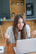 © simona - Young woman celebrates success while using a laptop at a modern kitchen table, showing excitement and engagement during remote work or video call in a bright home office setting.