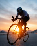 Cyclist rides at sunset, silhouette against golden sky. Warm light glows behind wheels and frame. Rider leans forward, focused and determined. Road stretches ahead, calm and quiet