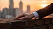 © TransPicHub - A person in business attire rests their hand on a brick wall with a city skyline blurred in the background during sunset.