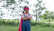 © Father_Studio - A confident young Asian girl smiles with arms crossed in a park. She wears a homemade pink superhero mask, cape, and denim overalls, showing girl power and imagination outdoors.