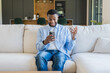 © Daniel - A young man sits on a light sofa at home, smiling and gesturing with excitement while looking at his smartphone during a video call, conveying connection, surprise and casual everyday life.