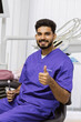 © Sofiia Shunkina - A smiling male dentist in purple scrubs gives a thumbs up while holding a dental mirror in a modern clinic