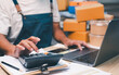 © waragron - Person wearing an apron uses a calculator and laptop while managing paperwork and packages, likely handling business or inventory tasks.
