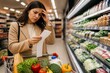 © mila103 - A woman stands in a supermarket with a shopping cart. She looks concerned as she reviews a grocery list. Fresh produce is visible on the shelves around her.