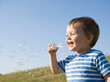 © Patcharin - Happy toddler boy blowing soap bubbles on a grassy hill under a clear blue sky, enjoying carefree outdoor play, laughter and a cheerful summer childhood moment.