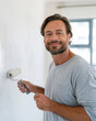 © lashkhidzetim - Man smiles while painting a wall with a roller. He wears a casual gray t-shirt. His expression shows satisfaction and ease. Natural light streams through a window behind him
