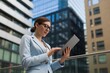 © Riyad Shikdar - Successful young businesswoman in suit using digital tablet standing outdoors against office buildings