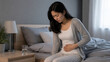 © Chanita - Pregnant woman sitting on bed edge, hand on abdomen, glass of water on nightstand, soft bedroom light, healthcare concept