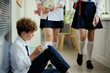 © DragonImages - Schoolboy sitting alone in hallway, writing in notebook while classmates passing by, conveying bullying, exclusion, loneliness. Useful for school counseling, anti bullying campaigns, education content