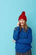 © SHOTPRIME STUDIO - A smiling woman wearing a blue knit sweater and red hat talks on a phone against a bright blue backdrop, casual cozy style, studio portrait for stock use today.
