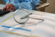 © Joi_studio - Close-up of financial documents being reviewed with a magnifying glass and calculator, auditing, compliance, financial analysis, and business verification in a professional office environment