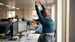 © Felippe Lopes - Young woman stretching arms up in joy and relief at her office desk with computers and colleagues in background