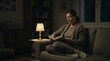 © Xenia - Woman reading a book in a dark living room illuminated by a single small lamp. Homeowner wearing warm clothes to save energy and utility costs. Financial struggle concept