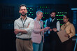 © Zamrznuti tonovi - It professional with crossed arms looking at camera, standing in a server room with a team discussing technology