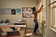 © Naseem - Teacher Decorating Classroom for Back to School | Smiling Female Educator Arranging Desk and Wall Art for First Day of New Elementary School Year.