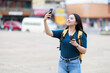 © Tj - Young Asian woman traveler holding a disposable coffee cup and smartphone, gracefully capturing a moment by taking a selfie or making a video call during her city exploration
