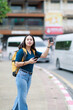© Tj - Young woman hailing taxi service with smartphone and coffee cup, backpack on shoulder, standing on sidewalk searching for ride in city street, urban travel lifestyle