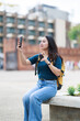 © Tj - Young Asian woman traveler sitting in a public square, holding a smartphone for recording or taking a selfie, and a coffee cup, creating travel content