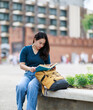 © Tj - Young Asian woman student enjoying reading a book while sitting on a wall with her backpack at Tha Phae Gate in Chiang Mai, blending education with travel lifestyle