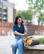 © Tj - Young female student smiling and multitasking, managing her studies on a laptop and conversing on a smartphone outdoors, representing modern education and connectivity