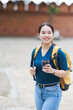 © Tj - Young woman exploring a historic city, carrying a yellow backpack and a takeaway coffee cup, happy and looking away with an old brick wall in the background