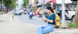 © Tj - Young asian woman traveler backpacker sitting on a curb by a busy street, looking at her smartphone and smiling, creating a relaxed moment amidst urban travel