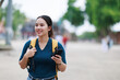 © Tj - Smiling young adult Asian woman with a backpack holding a smartphone, looking forward while walking outdoors, exploring new places during her travel adventure