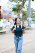 © Tj - Young woman smiling, walking on a city street, exploring and navigating with a smartphone and backpack, enjoying urban travel and student life outdoors