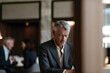 © Milos - A contemplative older man dressed in a suit appears lost in thought while sitting at a table, capturing the essence of reflection and focus in a modern business environment.