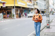 © Tj - Young Asian woman exploring city street, looking away while waiting, holding smartphone, wearing headphones and backpack, enjoying urban travel and journey discovery