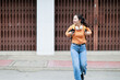 © Tj - Young Asian woman wearing an orange long-sleeve shirt. Jeans. A backpack. And white headphones around her neck. Walking confidently with a cheerful smile on a city street