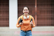 © Tj - Young Asian woman student carrying a backpack and headphones, smiling confidently while holding a smartphone, representing modern education and lifestyle