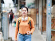 © Tj - Young Asian woman traveler walking in urban street, exploring city during solo trip, carrying backpack and headphones, enjoying journey and discovering new places