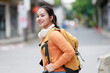 © Tj - Young Asian woman backpacker wears headphones around her neck while carrying a mustard-colored backpack. Smiling and looking back